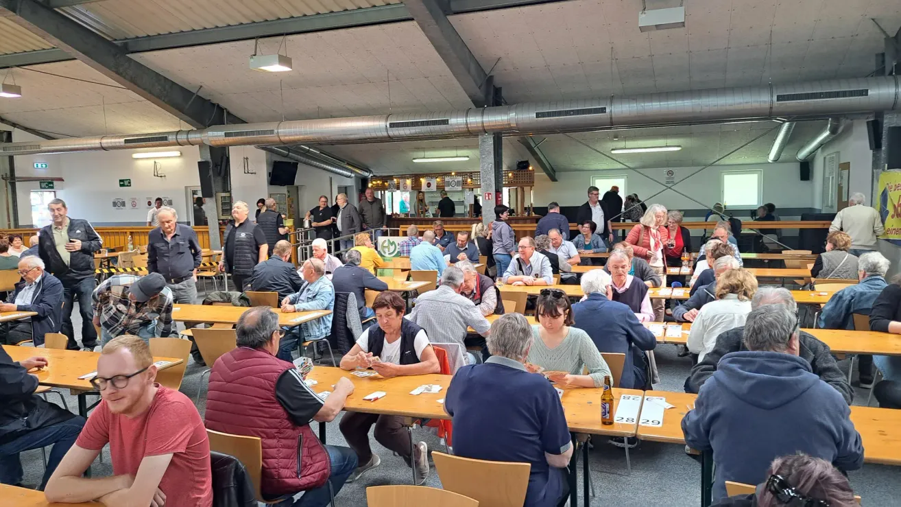 In a large indoor area, numerous individuals, including men and women, are seated at tables, engaging in card games. A few people are standing near the bar counter.