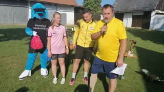 A man speaking into a microphone stands next to a girl and a mascot. They are on a grassy field with trophies and a building in the background.