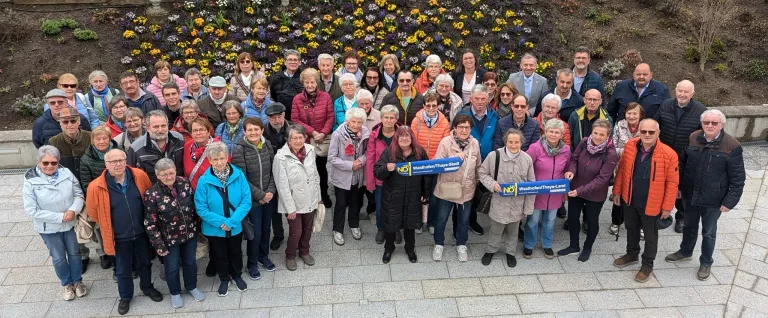 A group of elderly people stand together outside, holding signs and banners. They are posing for a photo in front of a wall adorned with flowers.