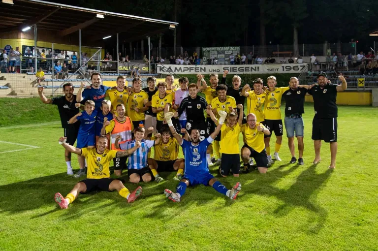 Fußballspieler posieren für ein Teamfoto auf einem Feld. Die Spieler tragen gelbe und blaue Trikots. Einige tragen Knieschützer. Dahinter jubelt das Publikum von den Rängen.
