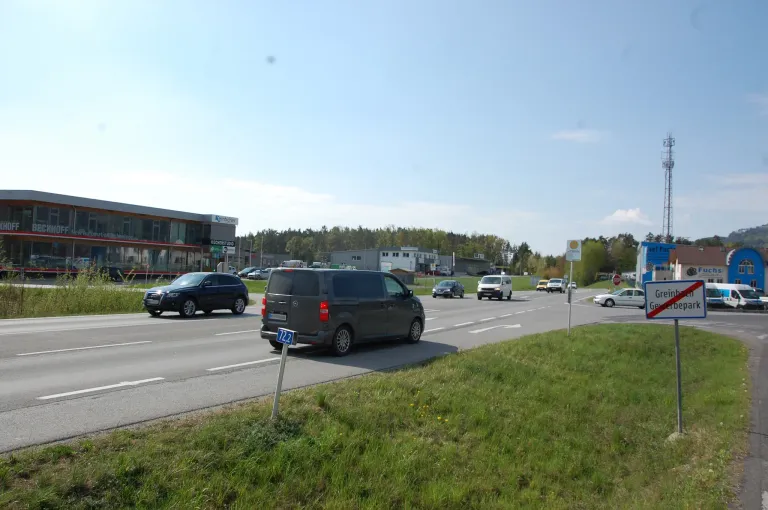 Eine belebte Straße mit mehreren Fahrzeugen, darunter ein dunkler Van mit der Nummer 72.2. Ein Stoppschild und Grasflächen sind am Straßenrand sichtbar.