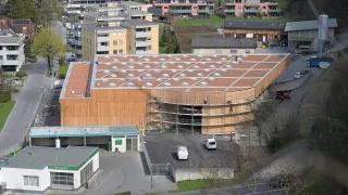 Luftaufnahme eines großen Holzgebäudes im Bau mit mehreren Arbeitern. Im Hintergrund sind zahlreiche Gebäude zu sehen, und eine Straße mit geparkten Fahrzeugen ist sichtbar.
