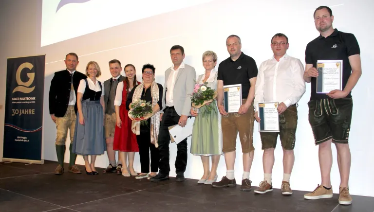 A group of people stand on a stage, holding certificates and flowers. They are dressed in traditional German attire, suggesting an award ceremony. They smile and look at the camera.