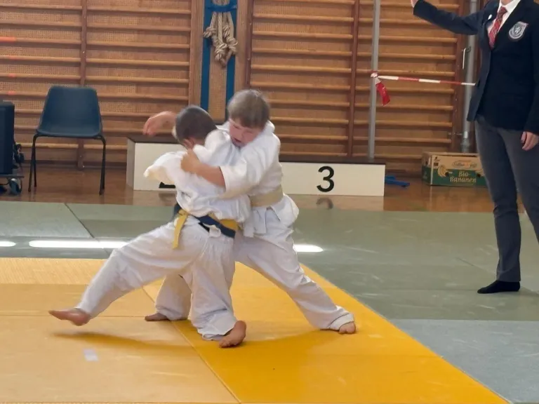 Zwei junge Jungen in Kampfsportuniformen ringen auf einem Teppich. Einer liegt auf dem Boden, der andere ist obenauf, beide konzentrieren sich auf den Kampf.