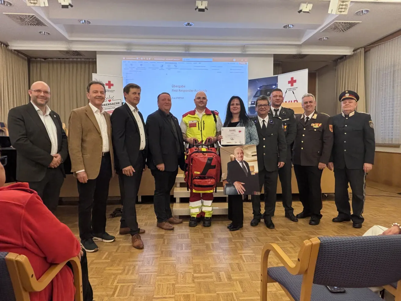 Eine Gruppe von Menschen steht in einem Raum, einige halten eine Urkunde. Ein Mann mit einem Rucksack steht vorne. Sie stehen vor einer Leinwand, wobei ein Mann in einer gelben Jacke ist.