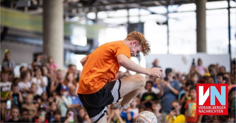 A young man in an orange shirt and black shorts performs a soccer kick in an indoor arena, with a blurred crowd watching.
