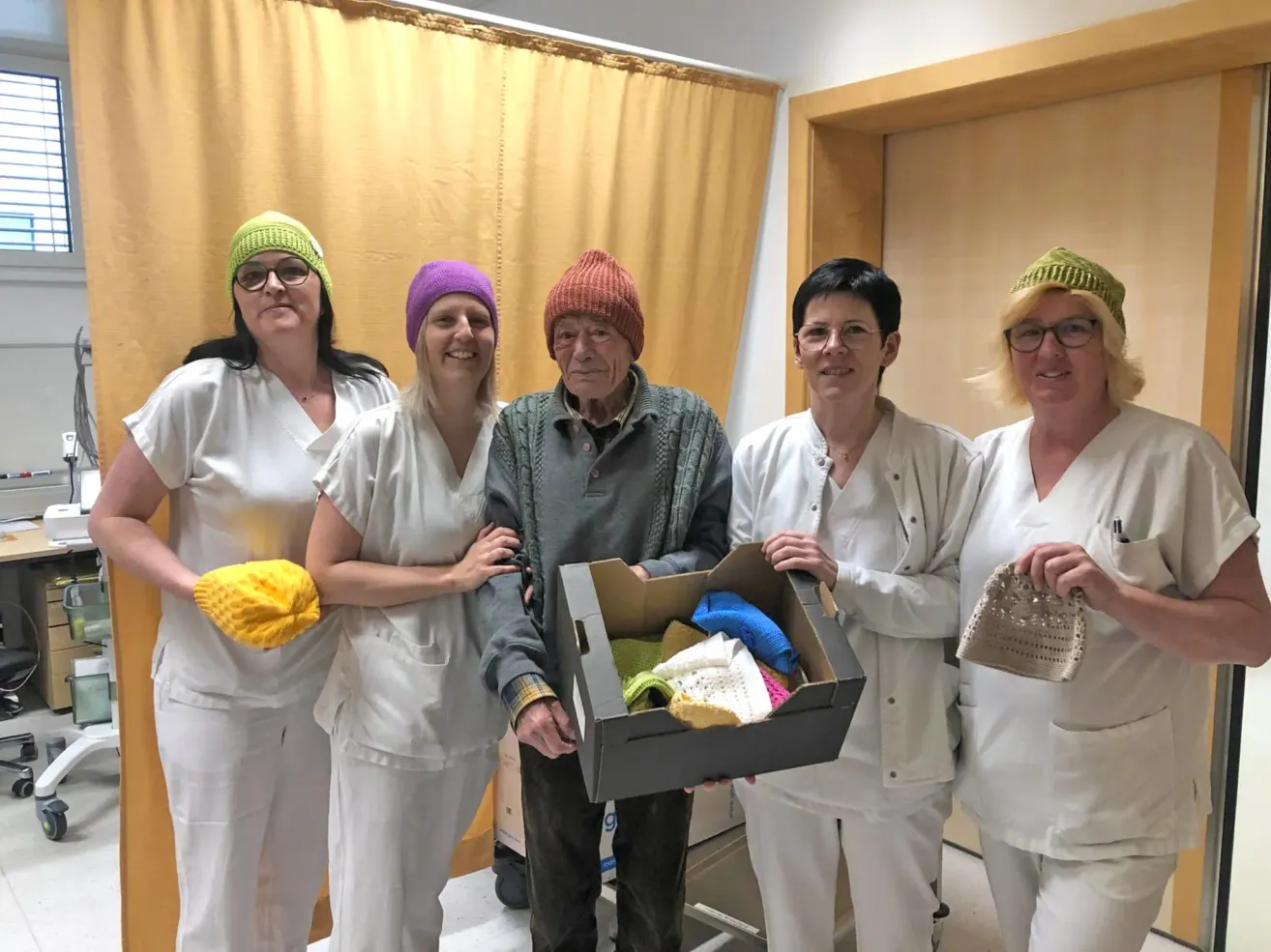Fünf Menschen in weißen Uniformen und Mützen stehen in einem Raum, einer hält eine Box mit gestrickten Gegenständen. Der Mann in der Mitte trägt eine Mütze. Sie alle lächeln für ein Foto.