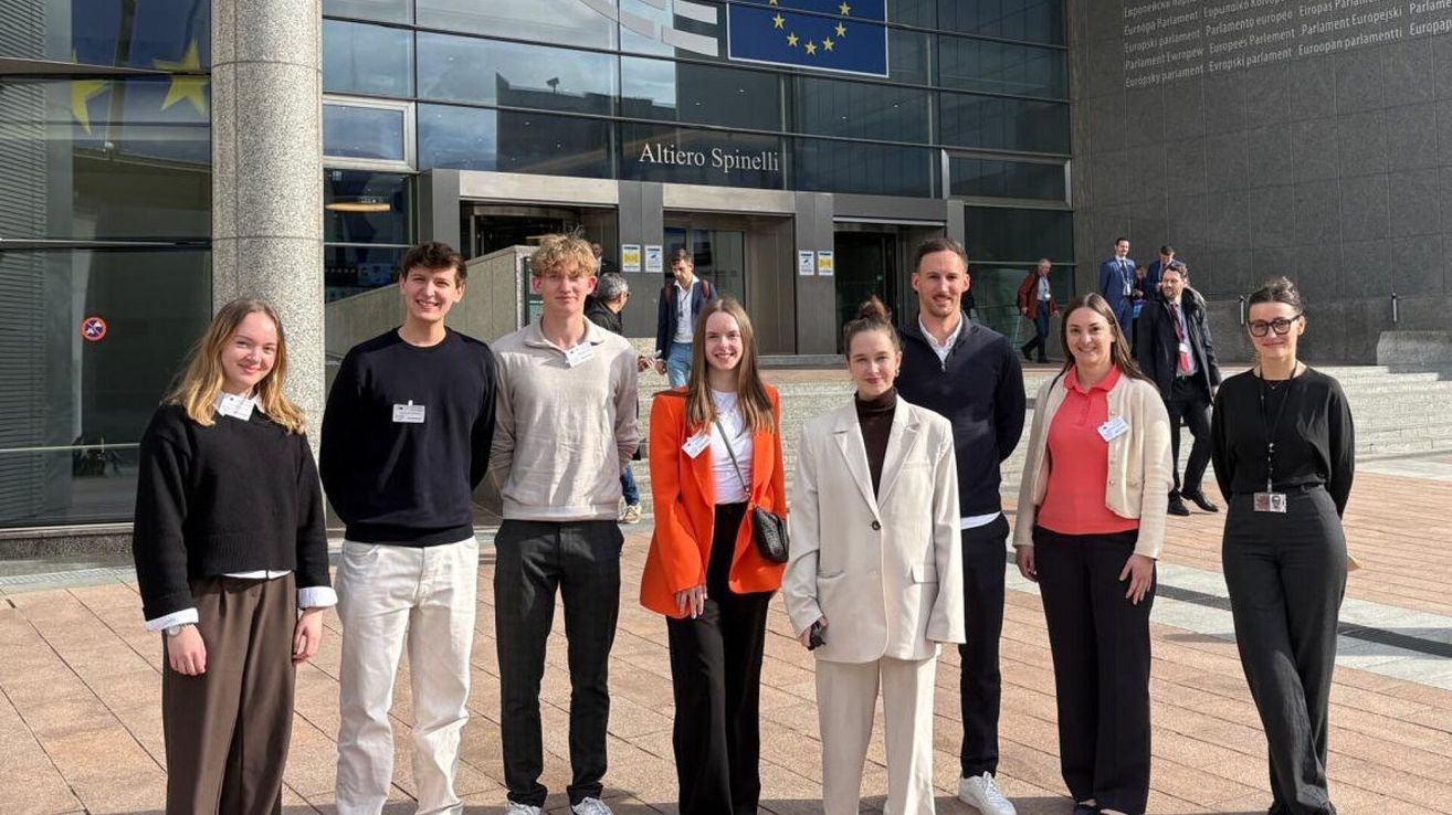 Eine Gruppe von Menschen steht vor einem Gebäude und lächelt für ein Foto. Das Gebäude zeigt die EU-Flagge und den Namen 'Altiero Spinelli'.