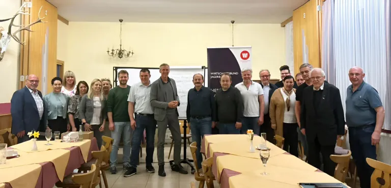 Eine Gruppe von Menschen steht in einem Raum mit einer Tafel und lächelt. Ein Tisch mit Weingläsern steht im Vordergrund.