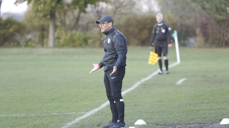 Ein Fußballtrainer ist auf dem Feld und gibt Anweisungen. Ein anderer Mann, möglicherweise ein Schiedsrichter, ist im Hintergrund.