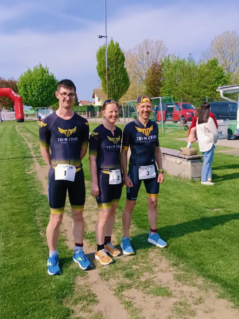 Drei Athleten in Radtrikots stehen auf einem Feld mit den Nummern 1, 2 und 3 auf ihren Trikots. Sie posieren für ein Foto, mit einer Frau im Hintergrund in der Nähe einer Betonbank.