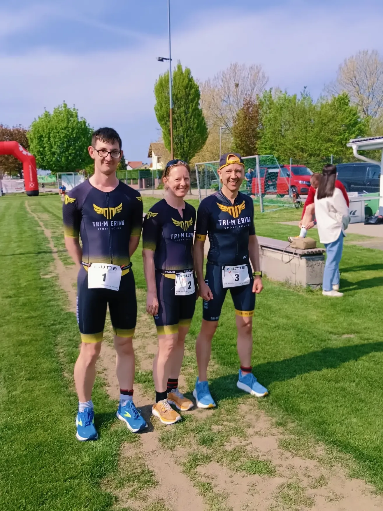Drei Athleten in Radtrikots stehen auf einem Feld mit den Nummern 1, 2 und 3 auf ihren Trikots. Sie posieren für ein Foto, mit einer Frau im Hintergrund in der Nähe einer Betonbank.