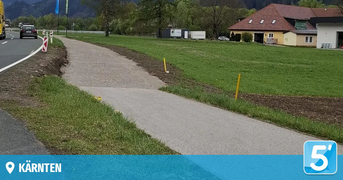 Ein Kiesweg windet sich durch eine grüne Wiese. Gelbe Stangen markieren den Weg. Ein gelbes Haus und ein geparktes Auto sind im Hintergrund.
