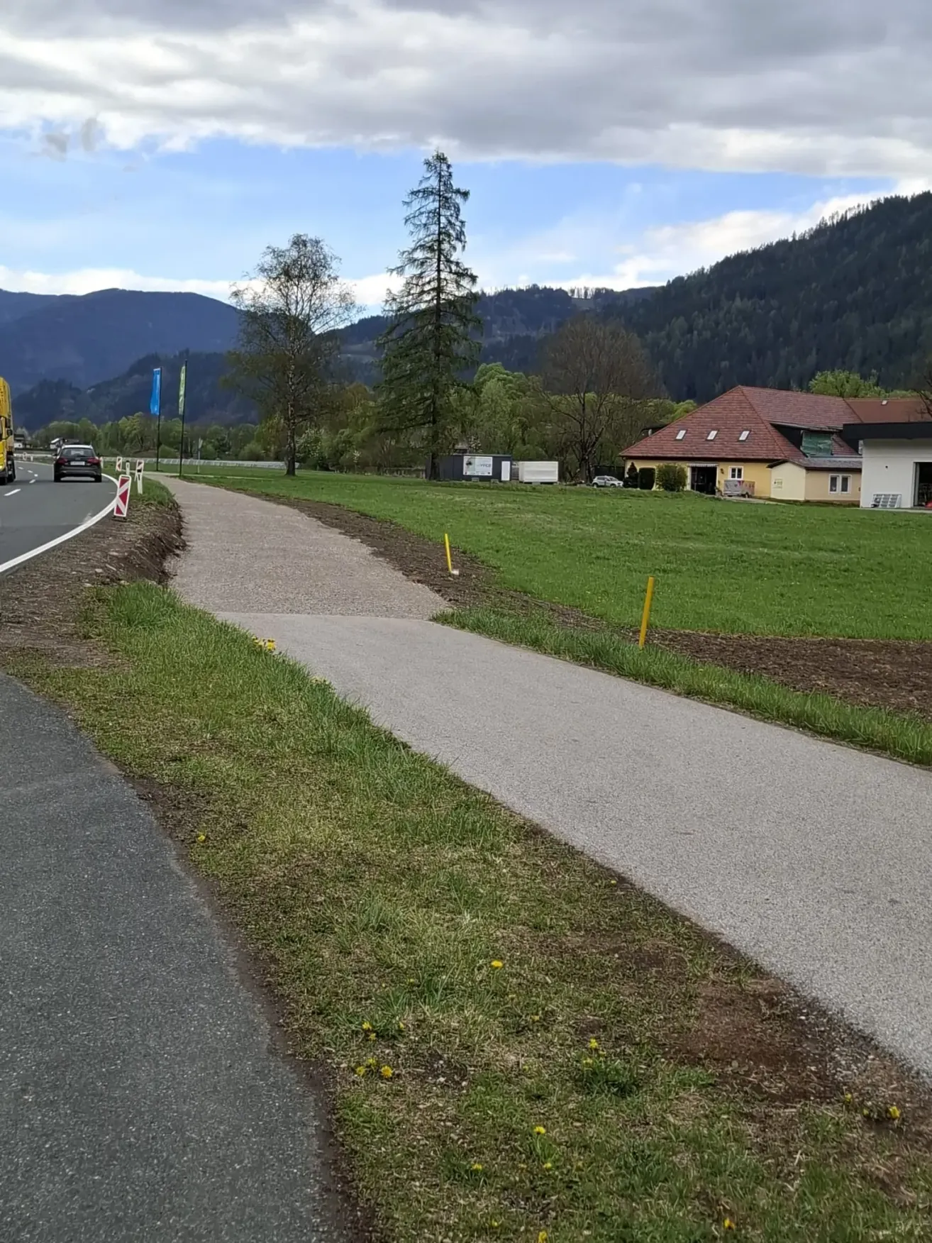 Ein Pfad neben einer Straße führt zu einem Haus mit einem braunen Dach. In der Nähe steht ein gelber Lastwagen und ein Auto. Das Gebiet ist von Bäumen und Bergen umgeben.
