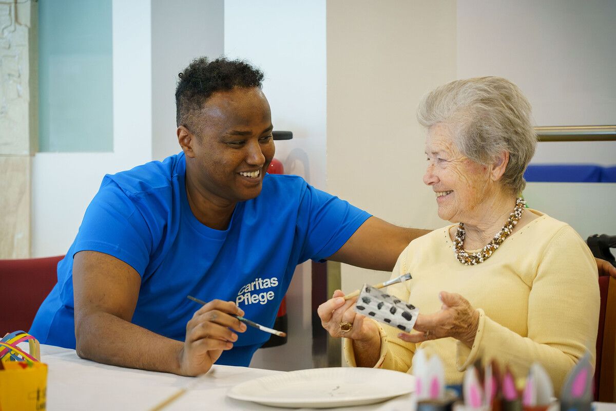 Eine ältere Frau lächelt, während sie eine Karte mit einem Mann malt, der ein Caritas Pflege T-Shirt trägt. Sie sitzen an einem Tisch mit Malutensilien.