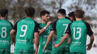 Fünf männliche Athleten stehen in einer Reihe. Sie tragen grüne Trikots mit Nummern auf dem Rücken. Sie scheinen miteinander zu sprechen.