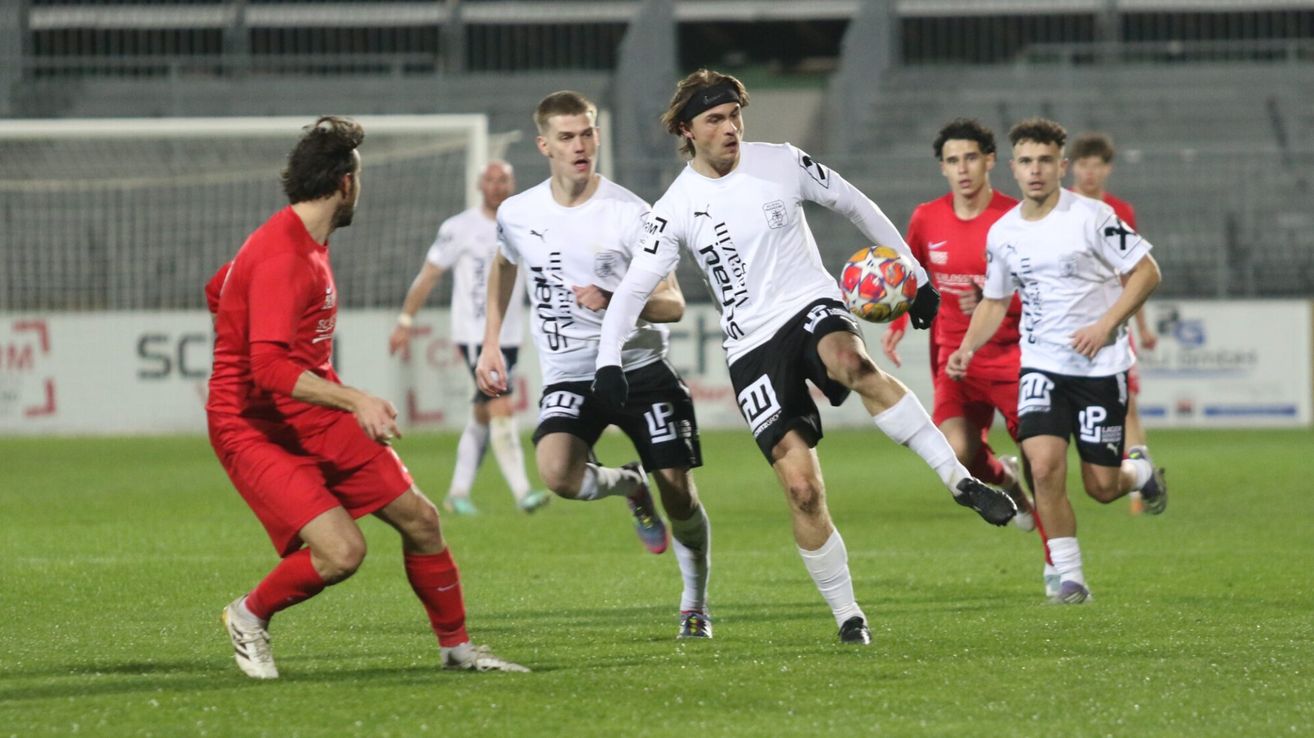 Fußballspieler in weißen und roten Trikots sind auf dem Feld. Der Spieler in Weiß tritt den Ball, während andere zusehen. Der Hintergrund ist verschwommen mit Tribünen und einem offenen Tor.