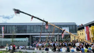 Eine große Menschenmenge versammelt sich um einen hohen Pfahl mit roten Bändern vor einem modernen Gebäude mit Glasfenstern. Viele sind in traditioneller deutscher Kleidung gekleidet und einige sitzen auf Bänken. Der Himmel ist bewölkt.