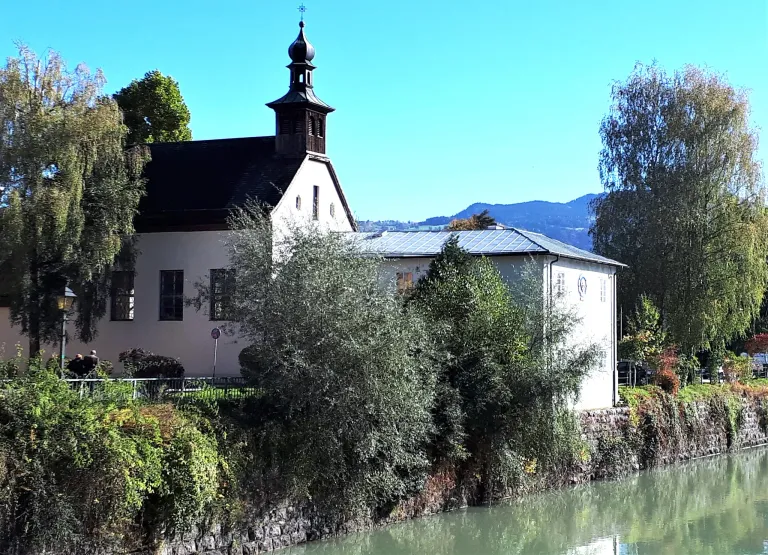 Eine Kirche mit schwarzem Dach steht am Fluss. Sie hat einen Turm mit einer Glocke und Solarpanele auf dem Dach. Im Hintergrund sind Berge sichtbar.