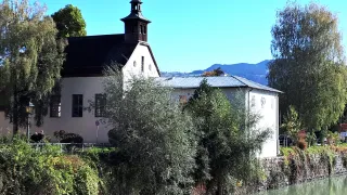 Eine Kirche mit schwarzem Dach steht am Fluss. Sie hat einen Turm mit einer Glocke und Solarpanele auf dem Dach. Im Hintergrund sind Berge sichtbar.