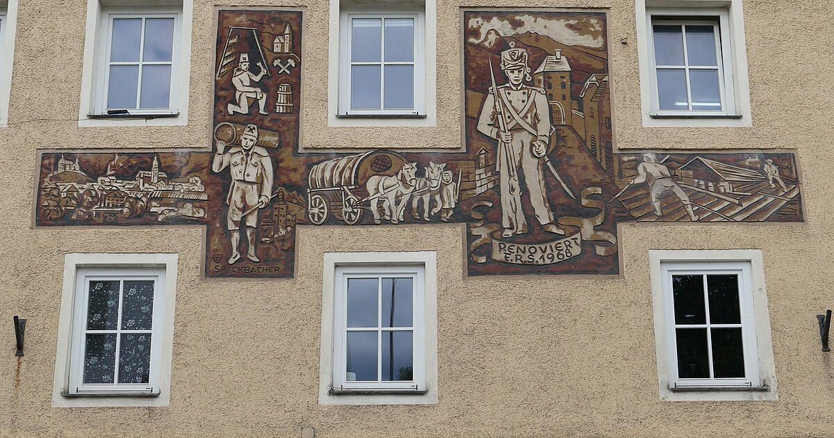 Ein Gebäudeaußenseite mit einem Wandbild, das einen Soldaten, einen Karren und einen Mann mit einem Fass zeigt. Das Wandbild hat einen Aufkleber 'RENOVIERT E.R.S.1968.' Zwei Fenster sind sichtbar.