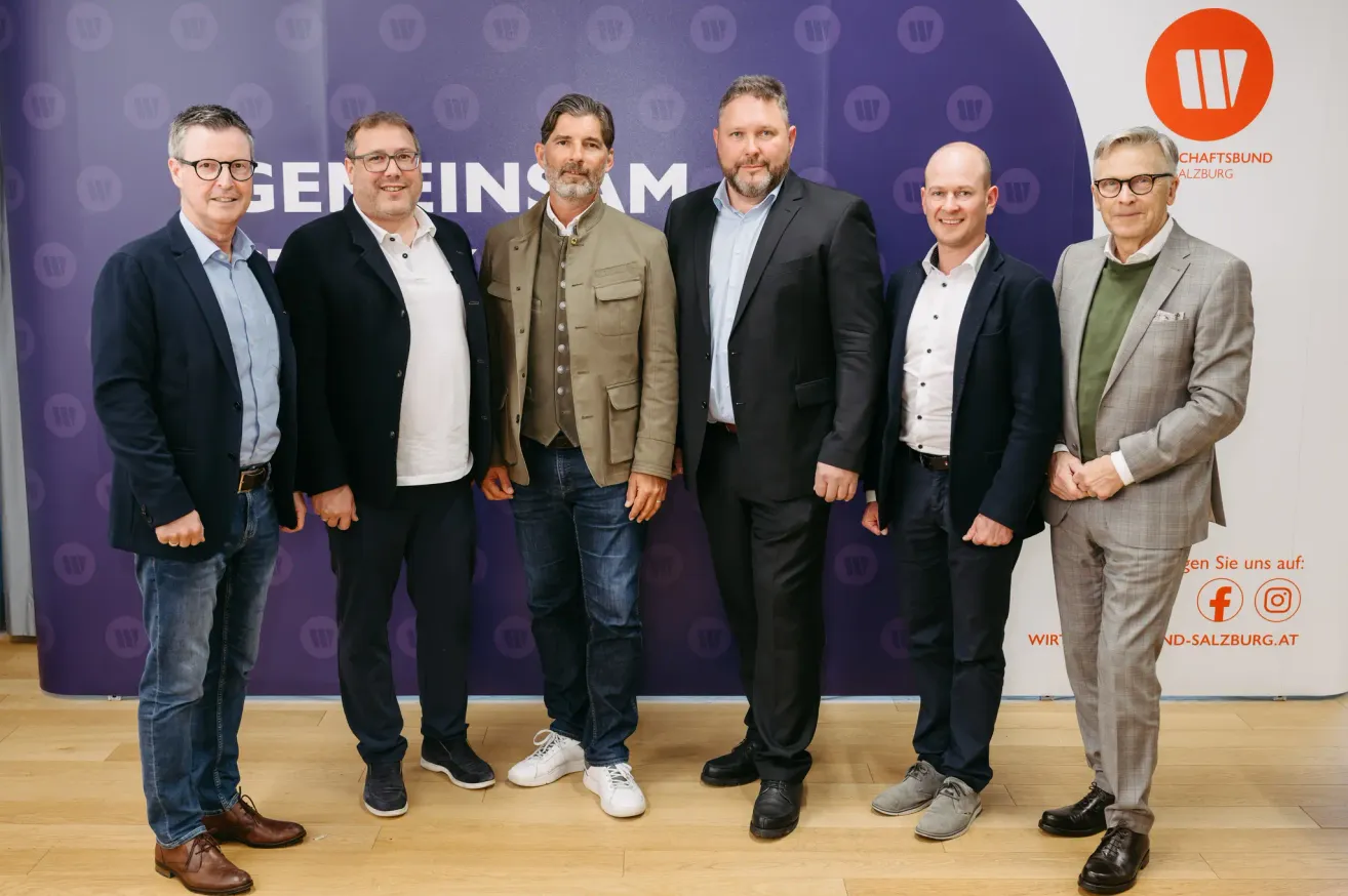 Sechs formell gekleidete Männer stehen vor einem lila Hintergrund mit weißem Text, lächeln und schauen in die Kamera. Sie scheinen sich bei einer geschäftlichen Veranstaltung zu befinden.