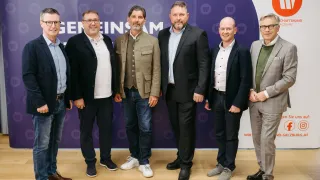 Sechs formell gekleidete Männer stehen vor einem lila Hintergrund mit weißem Text, lächeln und schauen in die Kamera. Sie scheinen sich bei einer geschäftlichen Veranstaltung zu befinden.