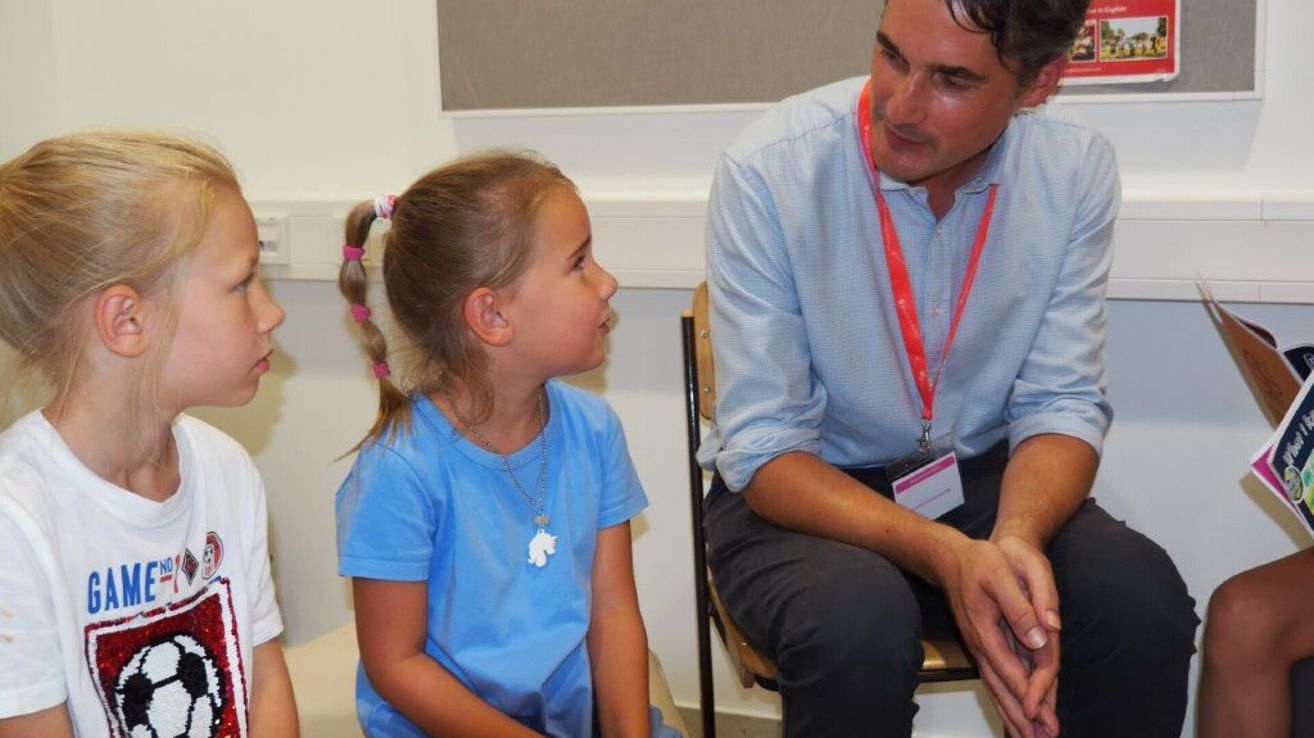 Ein Mann in einem blauen Hemd und einem roten Ausweisband sitzt auf einem Stuhl und unterhält sich mit einem kleinen Mädchen in einem hellblauen Hemd und Zöpfen.