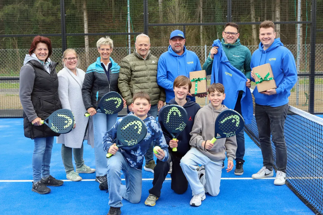 Eine Gruppe von Erwachsenen und Kindern posiert mit Tennisschlägern und Geschenken auf einem blauen Platz. Sie alle lächeln und einige halten Geschenke. Im Hintergrund befinden sich ein Zaun und Bäume.