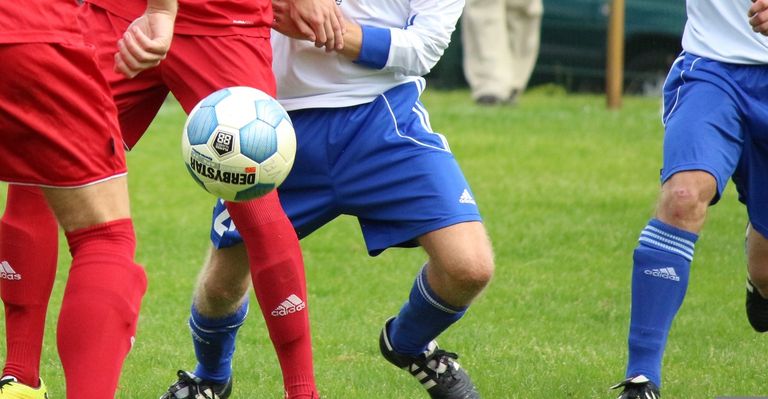 Two soccer players in red and blue jerseys are playing soccer on a grassy field. The player in red is holding the soccer ball, while the player in blue is attempting to kick it.