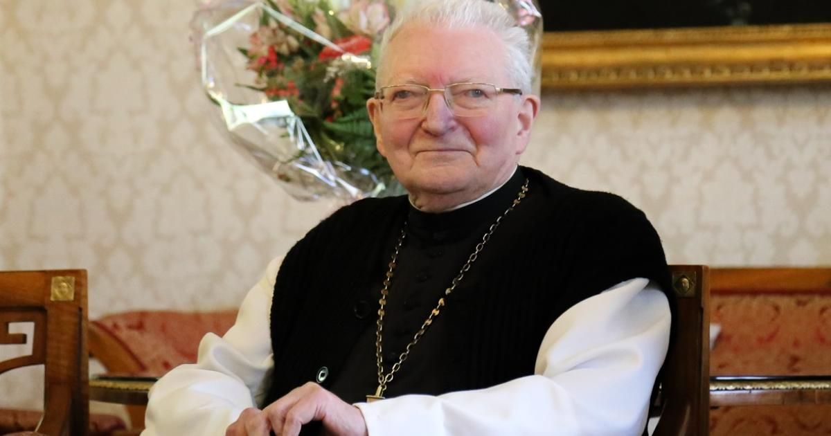 Ein älterer Mann mit einer Brille, einem schwarz-weißen Gewand und einer goldenen Kette sitzt. Ein Blumenarrangement steht hinter ihm.