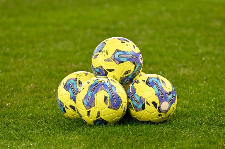 Vier gelbe Fußballbälle mit blauen und lila Designs sind auf einem grünen Feld gestapelt. Die Bälle werden wahrscheinlich für Training oder Freizeit verwendet.