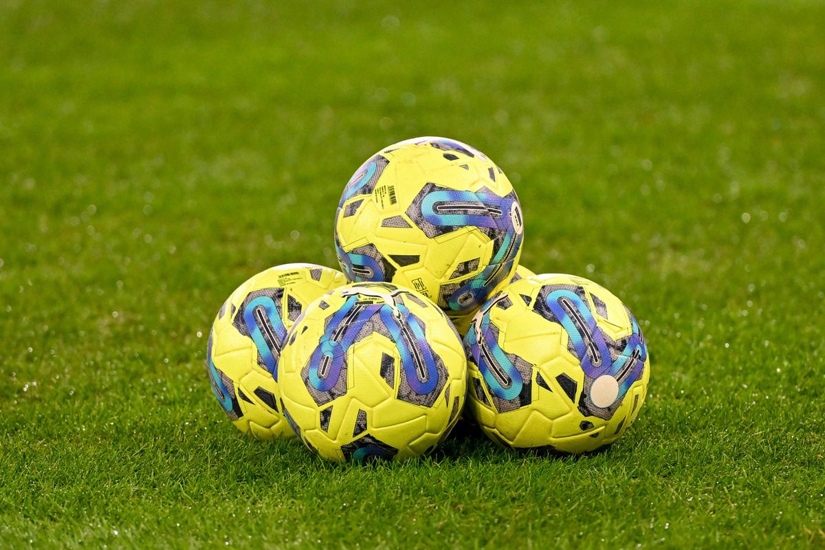 Vier gelbe Fußballbälle mit blauen und lila Designs sind auf einem grünen Feld gestapelt. Die Bälle werden wahrscheinlich für Training oder Freizeit verwendet.