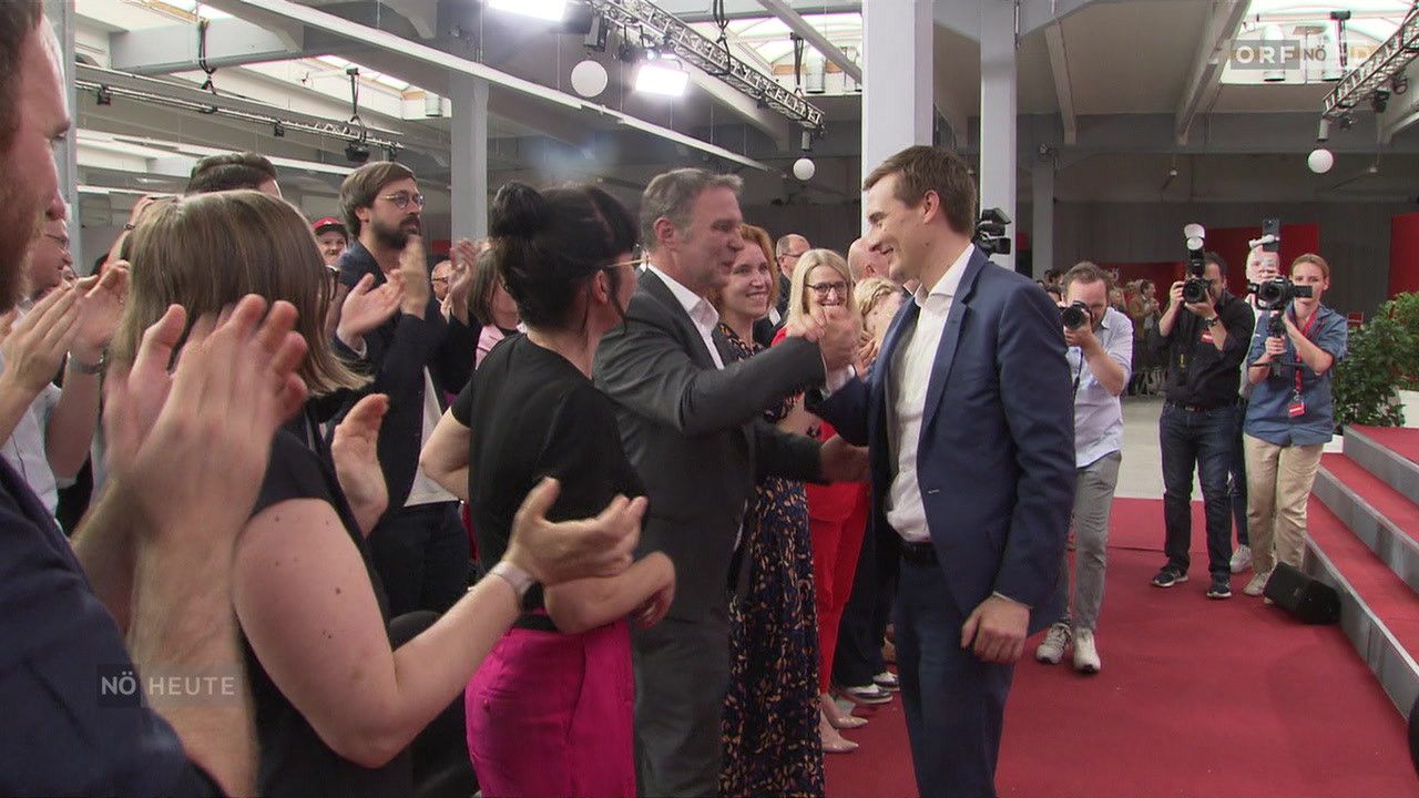 Zwei Männer schütteln sich die Hände in einem überfüllten Raum, während mehrere Leute klatschen und ein Fotograf im Hintergrund steht.