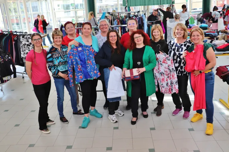 A group of women stand together in a mall, holding clothing and smiling at the camera. Behind them are clothing racks and other people shopping.