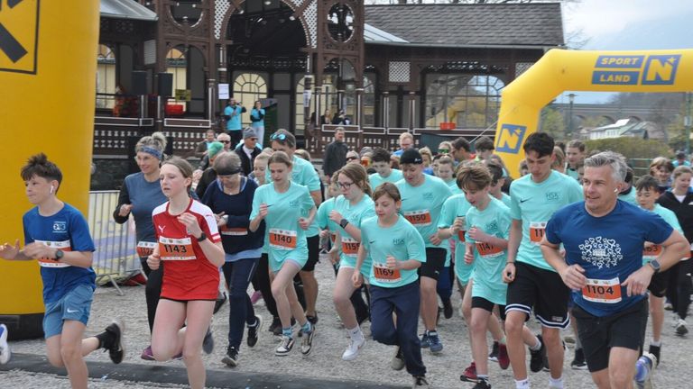 A group of young people in light blue shirts run in a race, with numbers on their shirts and a gazebo in the background. Spectators watch from behind.