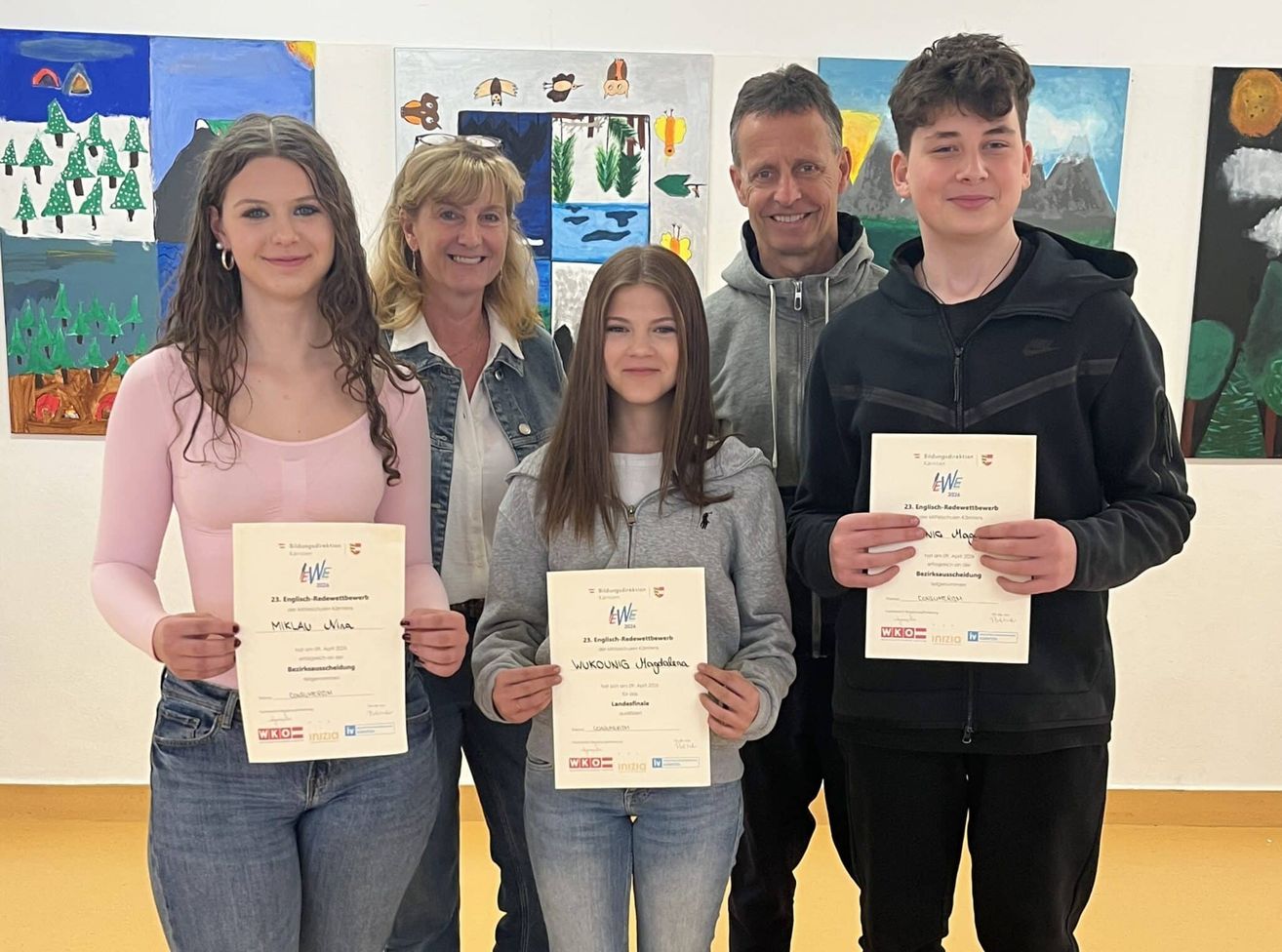 Five individuals stand together holding certificates. A woman in pink and a girl in gray hold certificates, while a woman in denim, a man in gray, and a boy in black also stand with them.