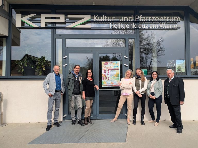 Sechs Personen stehen lächelnd vor einem Gebäude mit einer Glastür. Ein Schild an der Tür lautet 'Kultur- und Pfarrzentrum Heiligenkreuz am Waas'.