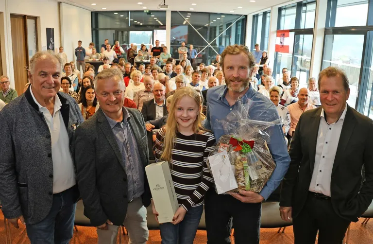 Drei Erwachsene und ein Kind stehen vor einer Menschenmenge in einem Raum. Der Mann links hält eine weiße Box. Der Mann in der Mitte hält einen Geschenkkorb. Das Mädchen rechts lächelt.