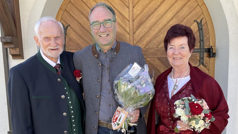 Three individuals stand close, smiling, with a man holding flowers. Behind them, a wooden door is visible. The man wears glasses and a gray jacket. The woman on the right has short hair, and the one on the left has a beard.