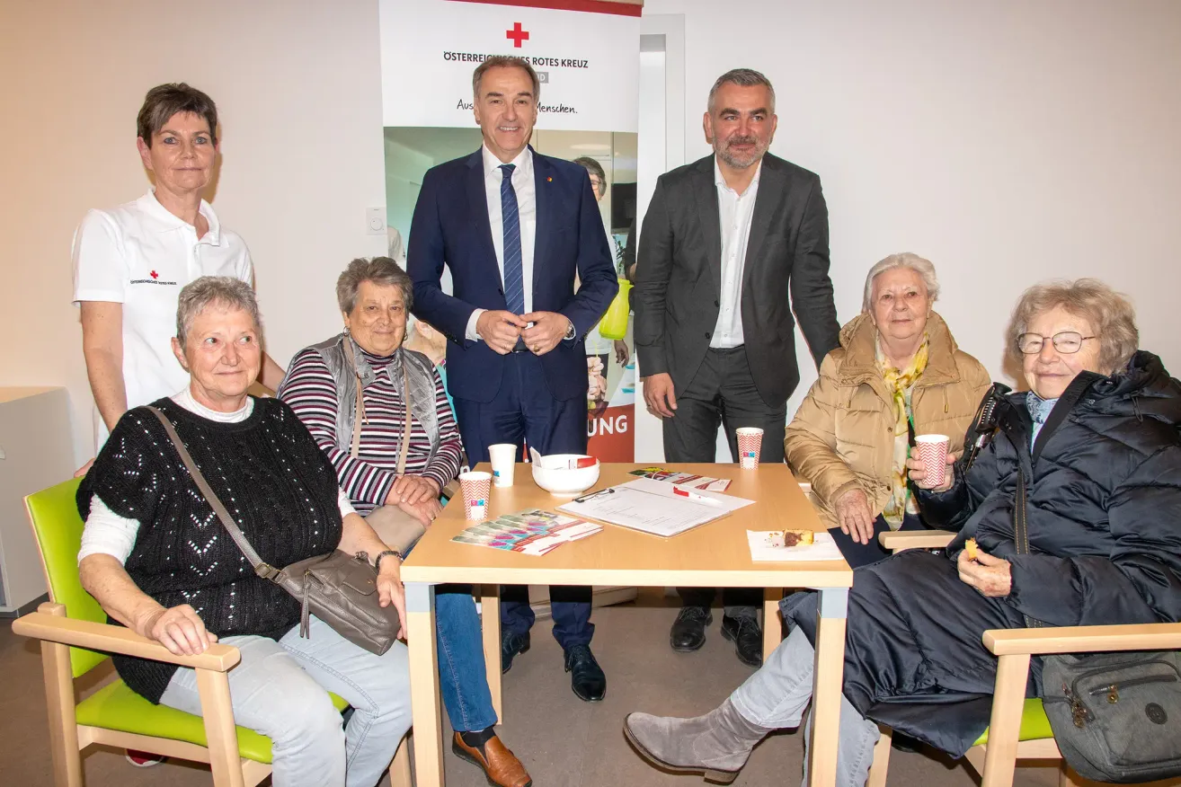 A group of people, including two men in suits, are gathered around a table with cups and papers, possibly for a meeting or discussion. A banner with the text 'Österreichisches Rotes Kreuz' is behind them.