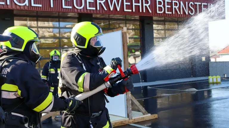 A firefighter practices using a fire extinguisher outside a building with the sign 'Feuerwehr'. Another firefighter stands behind him.