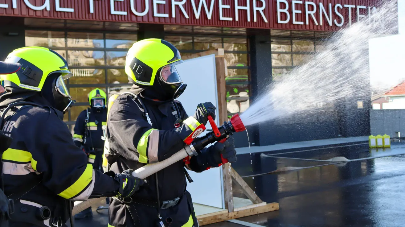 A firefighter practices using a fire extinguisher outside a building with the sign 'Feuerwehr'. Another firefighter stands behind him.