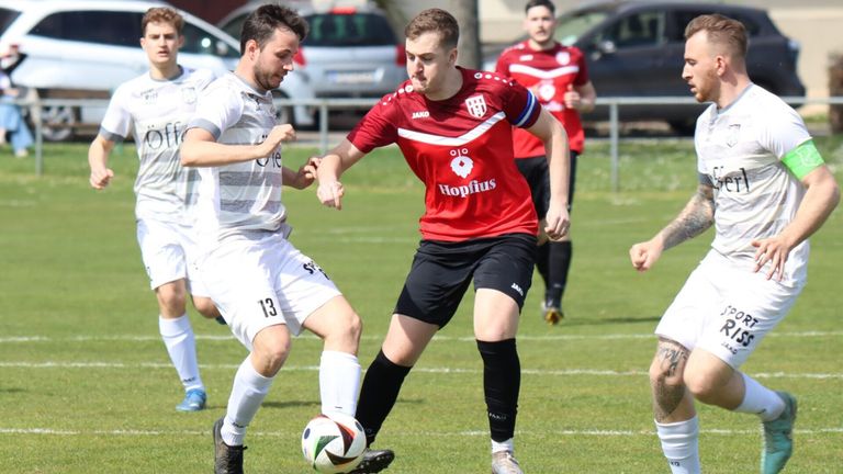 Zwei Fußballspieler spielen auf einem Feld. Einer trägt ein rotes Trikot mit dem Wort 'Hopfius', der andere ein weißes Trikot. Sie befinden sich in einer dynamischen Pose und kämpfen um den Ball.