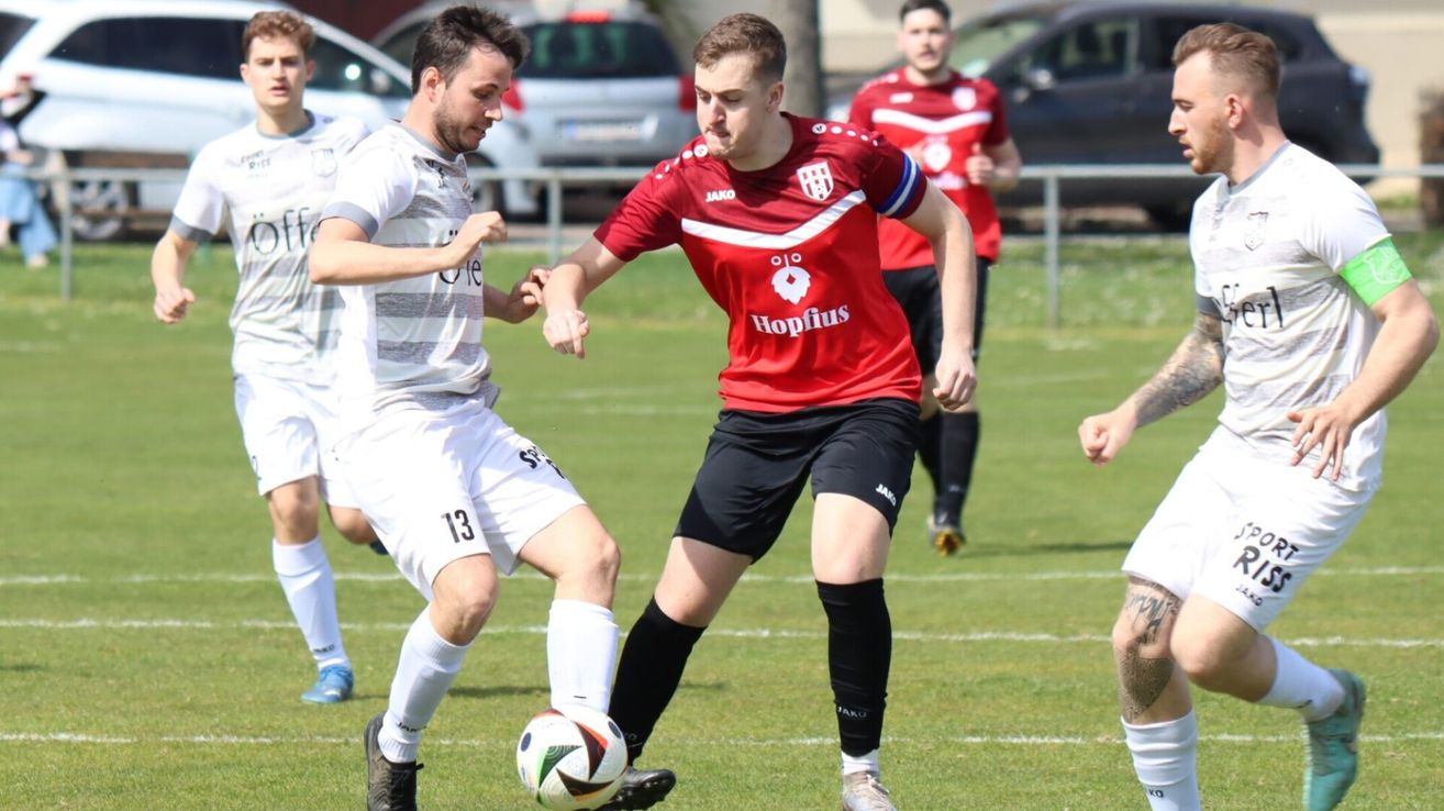 Zwei Fußballspieler spielen auf einem Feld. Einer trägt ein rotes Trikot mit dem Wort 'Hopfius', der andere ein weißes Trikot. Sie befinden sich in einer dynamischen Pose und kämpfen um den Ball.