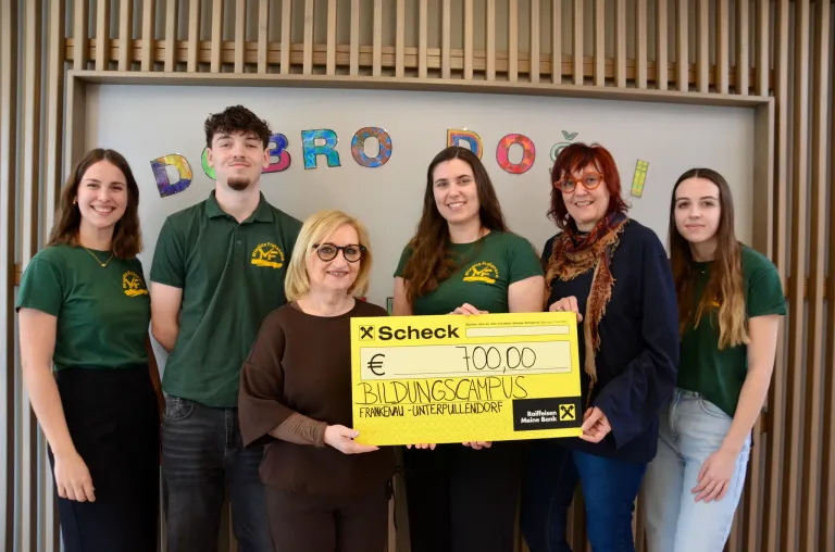 Five people stand before a white wall with colorful letters. A woman holds a yellow check for €700.00, with a woman wearing glasses and a scarf nearby. Behind them, a wooden wall and letters spell DOBROO.