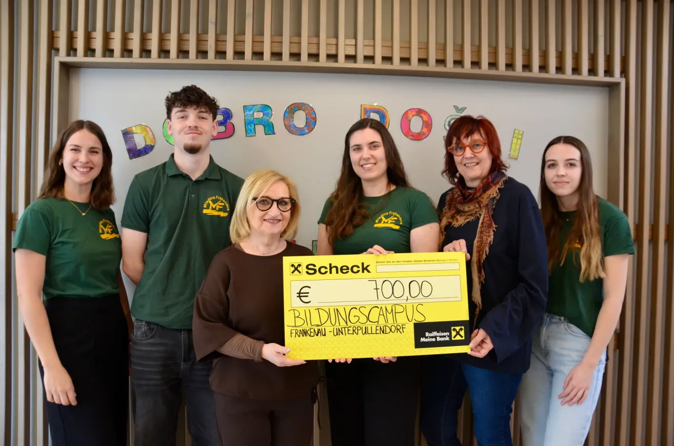 Five people stand before a white wall with colorful letters. A woman holds a yellow check for €700.00, with a woman wearing glasses and a scarf nearby. Behind them, a wooden wall and letters spell DOBROO.