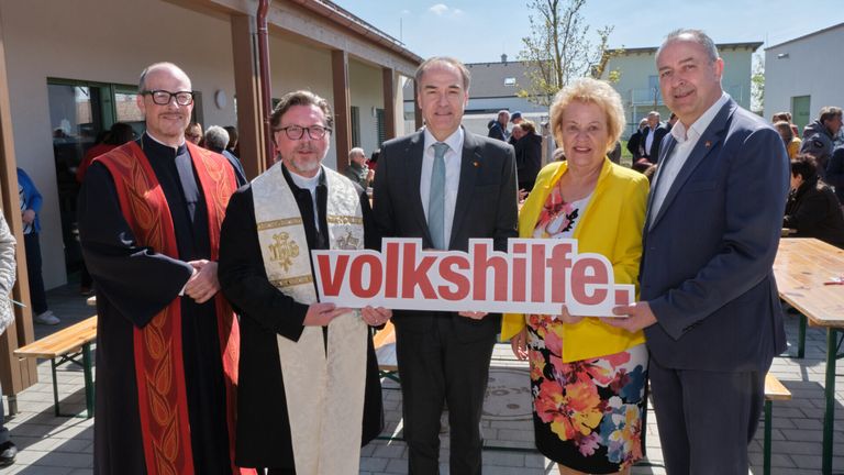 Several people stand in front of a building, holding a white sign with red letters that read 'volkshilfe'. They appear to be attending an event.