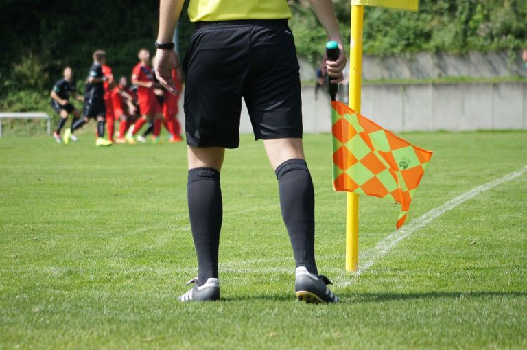 A soccer referee is standing on a green field, holding a whistle and a flag. Players in red jerseys are in the background.