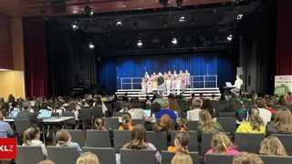 Eine Gruppe von Kindern in verschiedenen Kostümen steht auf der Bühne eines Theaters. Ein Erwachsener sitzt an einem Klavier auf der Bühne. Das Publikum sitzt in Reihen vor der Bühne.